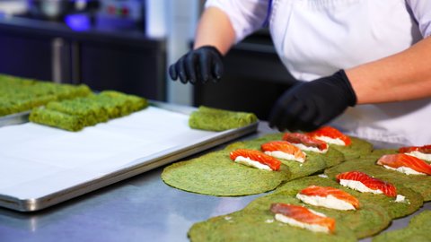 Unusual green crepes are rolled with cheese and salmon fish. The cook rolls the pancakes and lays them on the tray. Close up.
