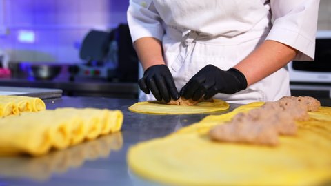 Careful hands of cook in gloves roll minced meat into a thin pancake. Ready-made crepes with stuffing at foreground. Close up.