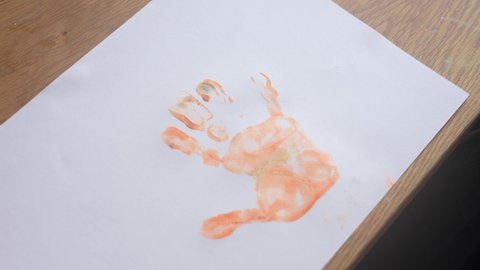 Top view 4k stock video footage of cute happy little baby boy making small handprints on white paper using orange paint 