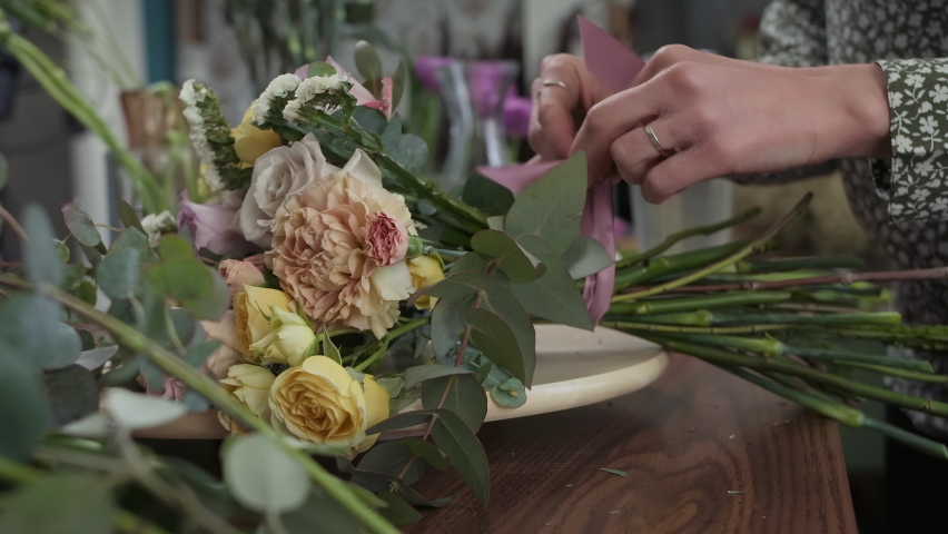 Professional woman florist preparing flower bouquet for sale or delivery. Female flower shop worker ties ribbon on bunch of roses lying on the table, close up | Shutterstock HD Video #1089380479