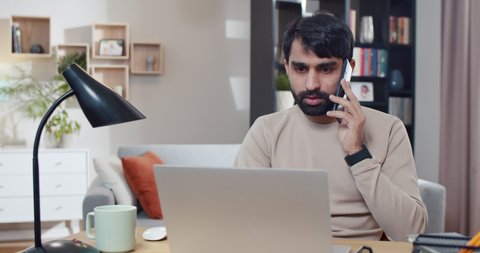 Young Arabian man speaking on mobile phone while working at laptop in cozy room. Arab businessman startupper talking on cellphone and sitting in front of computer screen at home. Freelancer.