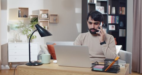 Young Arabian man speaking on mobile phone while working at laptop in cozy room. Arab businessman startupper talking on cellphone and sitting in front of computer at home. Freelancer. Conversation.