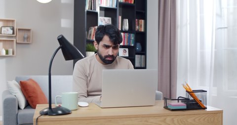 Young Arab tired and sad man working at laptop and getting bad news or result in job. At home. Arabian upset male freelancer at computer. Gamer loosing in game online. Lockdown concept. Lost money.