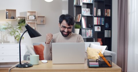 Young Arab handsome man in glasses working at laptop and getting good news or great result in work. At home. Arabian cheerful male freelancer sitting at computer. Gamer winning in game online.