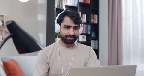 Close up of Arabian young handsome man in headphones sitting at table, listening to music and working on laptop. Arab male typing on keyboard of computer in nice cozy room. Freelancer in home office.