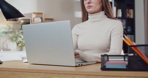 Pretty young Caucasian female tapping and typing on laptop at home. Pretty woman in glasses using computer and working. Freelancer on workday. Smart student studying online.