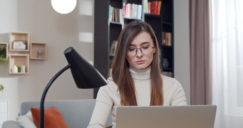 Young Caucasian woman in glasses sitting at desk in nice room and typing on laptop computer. Beautiful female freelancer working online and surfing the Internet. Tapping and searching info.