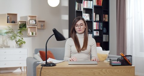 Caucasian young pretty woman in glasses sitting at desk, opening laptop computer and starting to work. Beautiful female freelancer start working day at home. Cozy room. Student studying online.