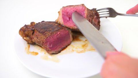 Macro closeup of hand cutting with fork and knife cooked rare blue red raw new york strip steak cut inside on white plate on table isolated against white background