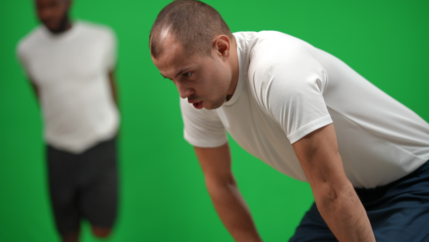Tired Caucasian sportsman resting as blurred African American man warming up at background. Multiethnic sports team of football players at chromakey green screen background