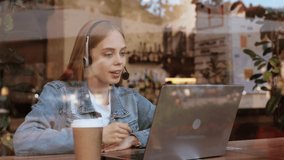 Young woman in headset looking at laptop camera and talking over video chat conference call recording vlog. Video call interview meeting. Portrait beautiful female freelancer speaking online in cafe