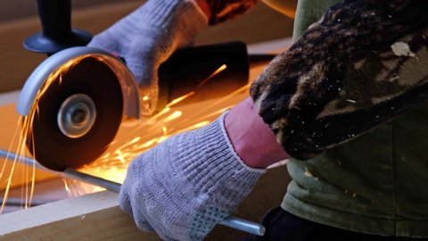 A builder's hands in work gloves cut a metal hairpin with an angle grinder electric tool. Preparation of a log for a wooden floor. Home repair with your own hands, a sawmill. Slow motion