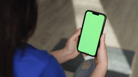 Unrecognizable woman hand holding smartphone on green screen chroma key background. Mobile phone mock-up for your product in vertical orientation portrait mode. Blank digital smartphone in woman hand.