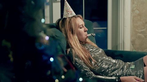 Side view of upset blonde girl lying in chic dress on couch or sofa and feeling lonely. Sad woman alone in party or birthday cap blowing whistle and celebrating birthday. festive cap and whistling