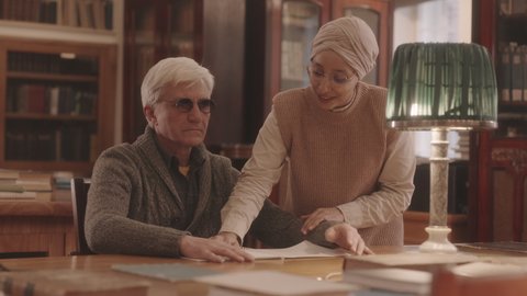 Medium slowmo of Caucasian senior man in eyeglasses reading Braille sitting by wooden desk at library. Muslim female librarian helping him to choose book