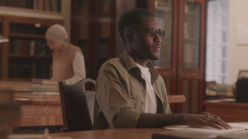 Medium slowmo of blind African American young man reading Braille book sitting by wooden desk at library