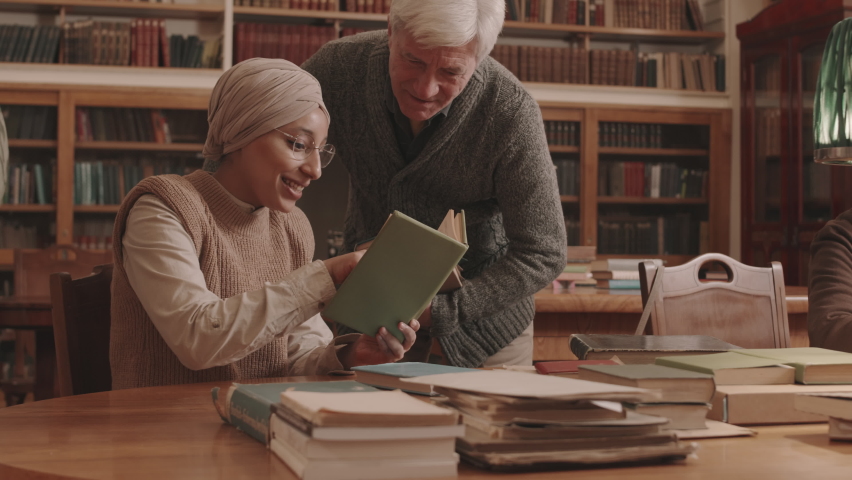 Medium slowmo of young Muslim woman in eyeglasses and headscarf and Caucasian old man discussing book plot at library