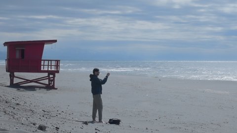 Travel blogger shoots panorama on empty beach with baywatch tower on background. Lonely young male traveler uses smartphone to create story for blog. Black sand coast, blue sky and amazing waves.