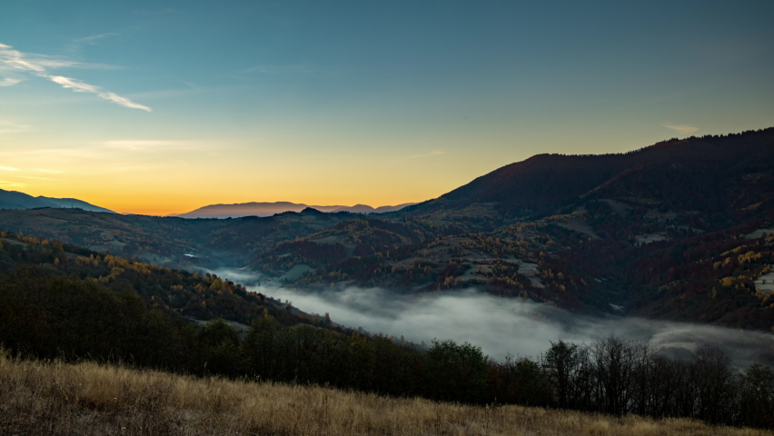 Sun rises in highland with big forestry mountains and morning fog fills valley among hill slopes, autumn under orange sky 4K time-lapse video