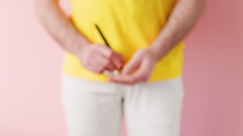 A man in white jeans writes on a piece of paper and shows a question mark drawn on it. Pink background. The concept of male diseases and impotence.