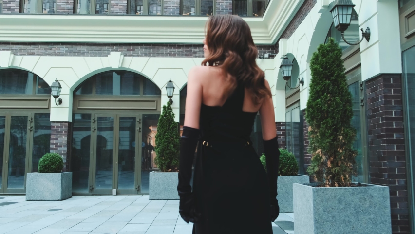 Sexy fashion model woman walking along city street gray sidewalk. Long loose brunette hair fly in wind motion. Wave curl hairstyle. Black evening trendy prom dress. Beauty strong face elegant makeup