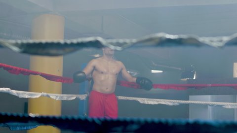 Young male boxer having break in ring corner, then doing shadow fight while training in boxing gym alone. Athletic man practicing punches and dodging techniques. Sport, boxing concept