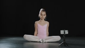 Teen girl dancer gymnast acrobat ballerina child making butterfly stretch exercise sitting on floor in dance studio hips stretching online video call remote distant class internet with phone on tripod