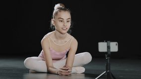 Caucasian girl child teen ballerina gymnast sitting on floor in dance hall waving hello to mobile phone camera online video call conference training with remote teacher using smartphone on tripod