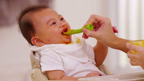 Side view mother feeding her asian baby daughter with pumpkin mashed or vegetable mash on rubber spoon.Mom trying to feed little baby boy at home enjoy and spending time together.Baby feeding Concept