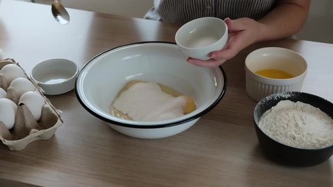 Woman adding salt in a bowl with eggs and sugar to beat the dough. The process of making dough for buns with ingredients on a table.