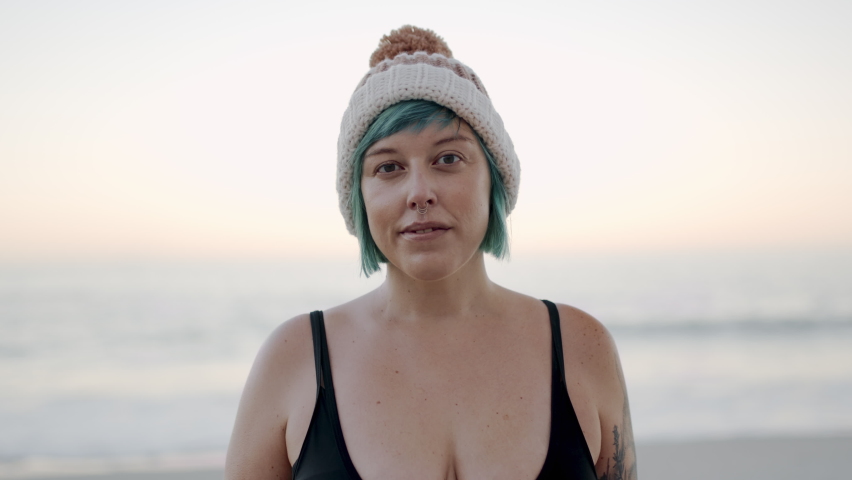Portrait of a woman in swimsuit wearing woolly hat standing on the beach. Beautiful female with short blue dyed hair and nose piercing looking at camera with a smile.