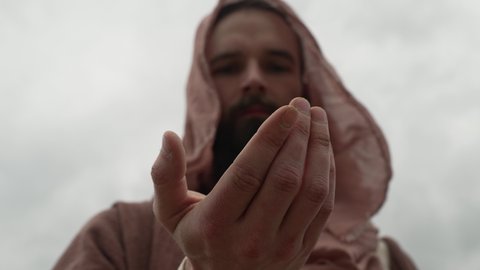 Jesus Christ or old testament bible prophet with a beard, wearing a brown robe and hood stands outside on cloud, overcast day holding and looking at rocks and dirt in his hand and then drops it.