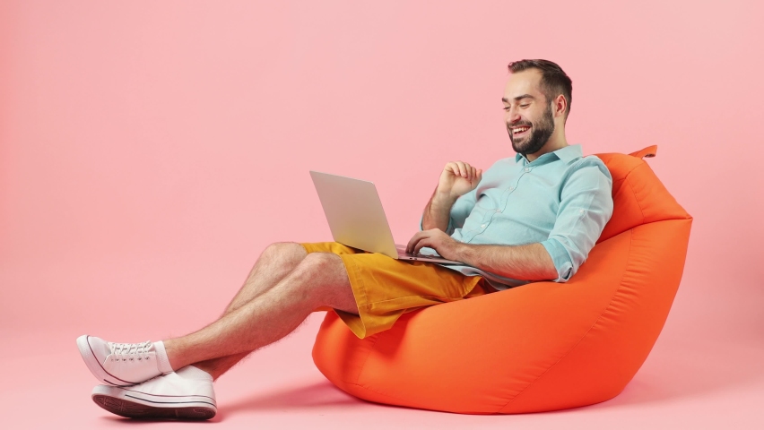 Full size body length happy fun young brunet man 20s wears casual clothes sit in bag chair hold use work on laptop pc computer doing video call isolated on plain plain pink background studio portrait