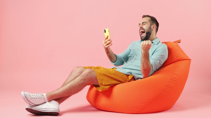 Full size body length fun young brunet man 20s wears casual clothes sit in bag chair hold use mobile cell phone say wow yes doing winner gesture isolated on plain plain pink background studio portrait
