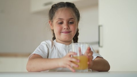 Girl sits at an empty table with a glass of orange juice. Healthy vitamin baby food. Kid with fruit drink in the kitchen. Funny kid drinks juice. Girl healthy food. Child eating breakfast in kitchen