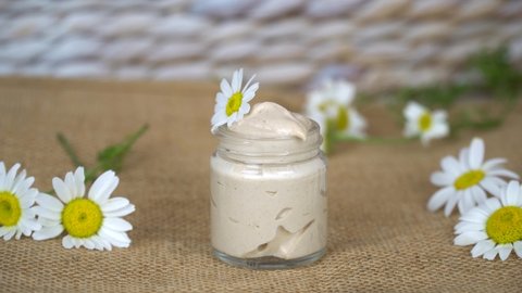 Homemade natural eye cream with chamomile flowers in transparent glass jar on beige background.