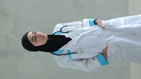 Vertical video of a female nurse. An Arab female doctor looks into the camera. Portrait of a Muslim woman doctor in a medical gown and black hijab, with a stethoscope hanging around her neck.
