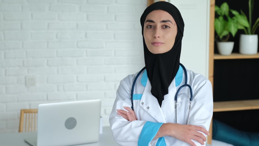 A friendly smiling female doctor in her office is waiting for new patients. A Muslim woman from the Middle East in a black hijab and medical coat works as a doctor in a private clinic. | Shutterstock HD Video #1089430969
