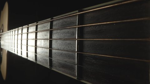 Camera pans over the fretboard of classical acoustic guitar against a black background. Classical guitar strings vibrate when playing a song. Brown wooden guitar neck with strings and frets close up.