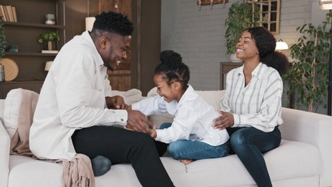Happy african family mum dad with cute little kid daughter tickling having fun relax on sofa together laughing young carefree parents and child girl laugh play cuddling at home on couch casual game
