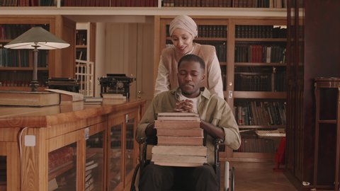 Tracking medium slowmo of young African American man with pile of books on his laps sitting in wheelchair carried by helpful Muslim female librarian towards wooden desk