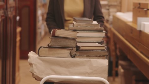 Midsection slowmo of unrecognizable female librarian carrying old rare books in cart while walking along drawers with card catalog in library