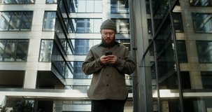 A young man with a beard, dressed in casual clothes and a knitted hat, smiles while typing on a mobile phone, standing in the courtyard of a modern glass building. Video in 4k, red komodo