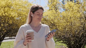Smiling pretty woman using mobile phone tapping scrolling screen of smartphone browsing internet social media app data holding hot coffee in the park near a tree with yellow leaves on warm sunny day.