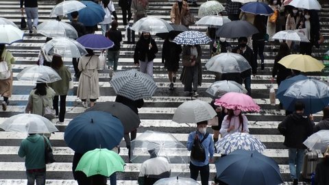 OSAKA, JAPAN - APRIL 2021 : Aerial top view of crowd of people wearing surgical masks to protect from Coronavirus (COVID-19) at the street. Slow motion shot in rain. Commuters walking with umbrella.