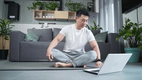 Happy Asian practices yoga at home, sitting in lotus position, man uses laptop for online classes, leads an active lifestyle doing exercise. watch lessons notebook computer learning