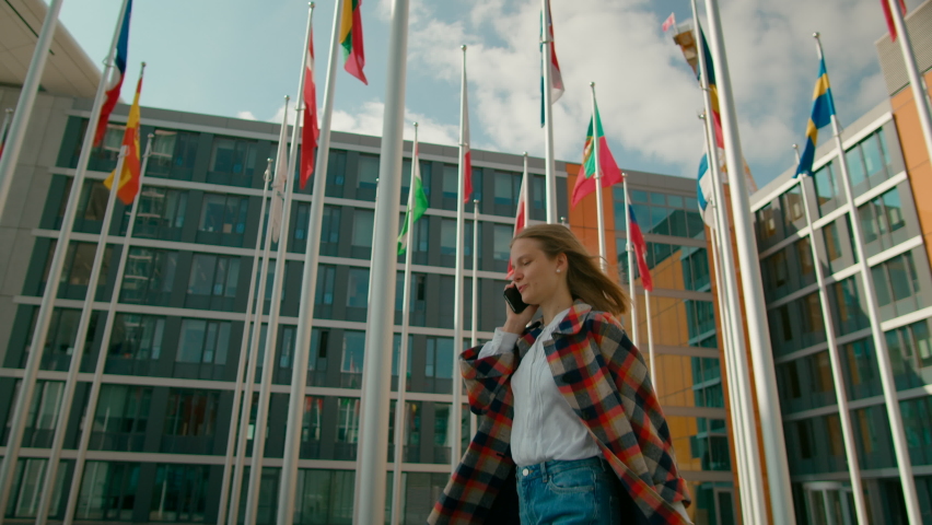Young Woman Walks and Talks by Phone in European Parliament administrative office Courtyard with Waving Flags of EU Countries in Luxembourg City. International Communications. 4K tracking shot Royalty-Free Stock Footage #1089438665