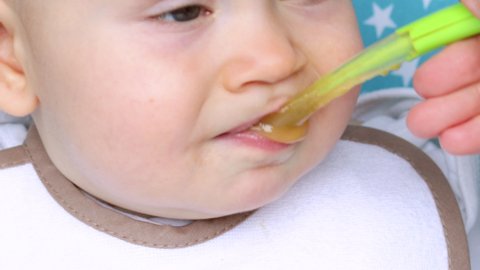 very close up video of a baby boy that is feed by mother or babysitter. cute toddler with beautiful big eyes. baby is eating mashed vegetables or fruit with green spoon. food diversification concept.