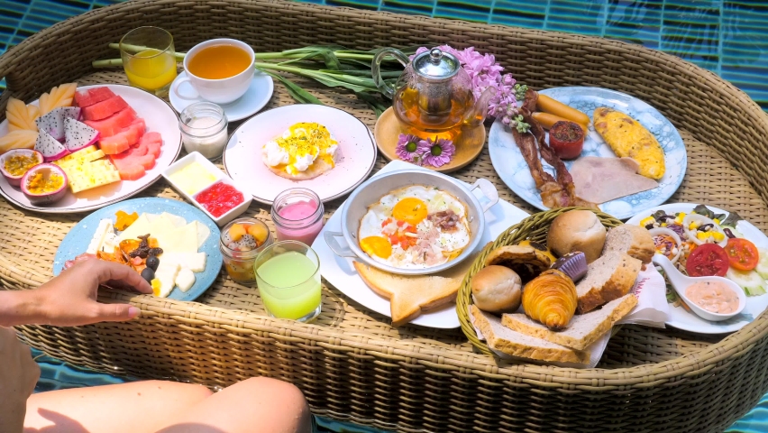 Woman hand take fruit juice glass of floating breakfast tray in swimming pool on hotel villa resort. Set with eggs benedict, sunny eggs, plate with tropical fruits, bread basket, tea cup and teapot. | Shutterstock HD Video #1089438833