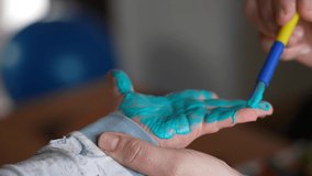 Mother or teacher preparing kid's hand to make hand prints on paper. Colourful child's hand covered with light blue paint. Slow motion 4k video